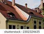 Small photo of Copper flashing detail on sloped red brown clay tile vintage roof. dormer. oval rococo style window. decorated stucco elevation. old Hungarian architecture concept. travel and tourism. metal flashing