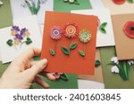 Small photo of Hand holding quilling card with flowers. woman making greeting cards. Hand made of paper quilling technique. Handicraft at home. Hobby, home office.