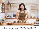 Small photo of Asian beautiful mature woman stay home, spend time in kitchen cooking. Young attractive carring female wear apron and smile, kneaded yeast dough with hand to baking bakery on table for dinner in house