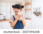 Portrait of Asian little cute kid holding a cup of milk in kitchen in house. Young preschool child girl or daughter stay home with smiling face, feel happy enjoy drinking milk and then look at camera.