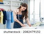 Small photo of Asian tailor woman talking on phone with customer in tailoring atelier. Attractive beautiful young female fashion designer working and checking new fashionable handmade clothes collection in workshop.
