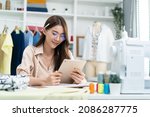 Small photo of Asian tailor woman working to design new clothes in tailoring atelier. Attractive young female fashion designer dressmaker use tablet to find reference to think and design new clothes in workshop room