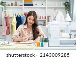 Small photo of Asian tailor woman working to design new clothes in tailoring atelier. Attractive young female fashion designer dressmaker create new pieces of clothing from patterns and designs in workshop room.