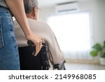 Small photo of Asian daughter support old disabled man sitting on wheelchair at home. Beautiful girl help and take care of senior elderly mature handicap father patient doing physical therapy in living room in house