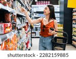 Small photo of Asian young beautiful woman holding grocery basket walk in supermarket. Attractive girl feel happy and relax, enjoy picking up goods product from shelf in shopping department. Healthy shopping concept