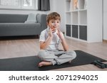 Small photo of A child boy sits on a black sports mat at home on the living room floor, smiles and spits into the camera. Recoil between exercise or yoga poses