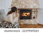 Young woman sitting at home by the fireplace with a hot tea or coffee mug and warming her hands, she is wearing white woollen sweater. Cold houses in Europe concept during energy and gas crisis.