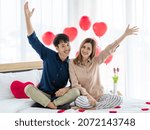 Happy Asian man and woman raising arms and looking at camera with smile while sitting on bed on Saint Valentine Day on bed in bedroom at home.