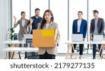 Small photo of Portrait shot of Asian sad jobless businesswoman in casual suit standing holding belongings in cardboard box after fired while male and female colleagues waving hands goodbye in blurred background.