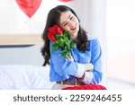 Millennial Asian young romantic cheerful female girlfriend sitting on bed in bedroom decorate with pink heart shape balloon holding red roses bouquet smiling on anniversary valentine day celebration.