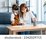 Small photo of Millennial Asian cheerful happy young beautiful female teenager mother nanny babysitter sitting smiling holding color pencils drawing painting together with little cute preschooler daughter girl.