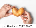 Hands holding heart-shaped potato above white background. Ugly food, funny vegetable concept. Horizontal orientation, top shot.