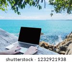 Close up hand typing laptop with blank screen on wooden table over summer beach background. Freelance working at anywhere and using social media communication concept