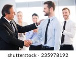 Good job! Two cheerful business men shaking hands while their colleagues applauding and smiling in the background