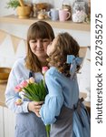 Small photo of Cute pretty little girl is greeting hew Mom with Women's or Mother's Day, giving her beautiful spring flowers and smiling. Mother and daughter at home, enjoying domestic life. Family values