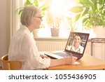 Senior woman in her sunny living room in front of a laptop making notes during a video call with her female doctor
