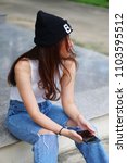 Small photo of A girl with white sleeveless, blue jeans, and black wool hat sitting on the floor, holding her phone with absent-mindedness