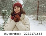 Portrait of happy young woman in outerwear
warming hands in mittens while standing in snowy weather in forest and looking at camera 