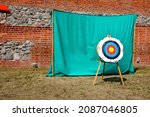 A view of a medieval shooting tange with a target in the shape of a round circle standing next to a green cloth situated next to the wall of a medieval castle or church seen on a sunny summer day