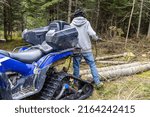 Small photo of Forestry worker is seen from the rear, standing behind a blue quad bike, holding towrope chain used to move cut down trees in woods. With copy space.