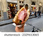 Small photo of Traditional Florentine street food Sandwich with Schiacciata bread, Salami slices and rocket salad in a hand on blurry background of a street in Florence, Italy