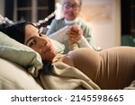 Small photo of Smiling pregnant woman and midwife at home. Woman in casual clothes lying on bed, Asian doula holding hand. Pregnancy, medicine, home birth concept