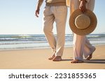 Middle-aged couple enjoying vacation at seashore with beautiful seascape in background. Legs closeup of lady with straw hat in hand and man strolling barefoot on wet sand. Romance, holiday concept