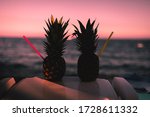 Small photo of a pair of cocktail in a fresh pineapple fruit with a swizzle stick against the sea during sunset. Tropical summer vacation concept. Pink sky during sunset on the beach of a tropical island