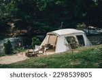 Small photo of Camping in a white tent next to water with camping chairs and lanterns in woods and trees with sunlights during sunset in golden hour.
