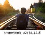 A boy with a backpack on his back sits on train tracks at dusk 