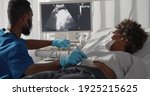 Small photo of Competent afro male doctor in medical protective mask doing ultrasound screening for pregnant woman at modern clinic. Young afro woman in safety mask visiting gynecologist