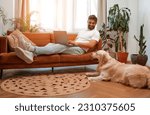 Small photo of Handsome bearded man sitting on sofa by window with laptop along with labrador dog lying on floor while freelancing online, shopping or chatting in living room at home.