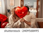 Happy Valentine's Day. Young couple in love holding a heart-shaped balloon, covering themselves with it while kissing, sitting on the sofa in the living room at home. Romantic evening together.