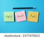 Small photo of Paper on blue background with pencil writing bully words UGLY FAT IDIOT, concept of someone who hurts or frightens victims by degrade or demean in some way to feel powerful or stronger