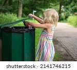 A toddler girl in a dress with colored stripes throws a plastic bottle into a green garbage can in a park.