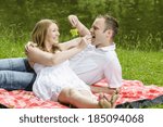 Young couple sitting together in a summer park and have a picnic on a checkered picnic blanket. They feed each other with treats.