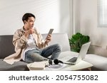 Small photo of Happy smiling Asian student sits and relaxes on a couch using modern tablet browsing unlimited wireless internet, positive young man freelancer working uses wifi at a home apartment, technology idea.