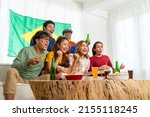 Small photo of Group of Asian people friends sit on sofa watching soccer games competition on TV with eating food together at home. Man and woman sport fans shouting and celebrating sport team victory sports match