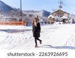 Young Asian woman in black coat walking on small town street covered in snow in snowy day. Attractive girl enjoy travel local village near the mountain in Japan on holiday vacation in winter season.