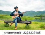 Young Asian man relax and enjoy outdoor lifestyle travel nature camping in forest mountain on summer holiday vacation. Handsome guy drinking coffee and playing guitar by the lake in the morning.