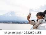 Happy Asian woman enjoy and fun outdoor activity lifestyle using mobile phone taking selfie with Mt Fuji covered in snow during travel nature road trip on car at Kawaguchi lake on holiday vacation.