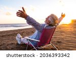 One mature and old man enjoying the summer vacations alone at the beach sitting in a small chair looking to the sea. Male person feeling free with opened arms. Freedom concept.