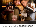 Small photo of Two glasses stands on bar and two young bartenders fills them with smoke and alcohol at the same time