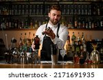 Small photo of various steel shakers and bottles stand on the bar counter, and man bartender gently pours an alcoholic drink from jigger in the shaker cup