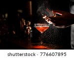 Bartender in black apron and blue shirt sprays an orange peel in cocktail glass with ice at a bar counter