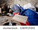Small photo of Houseless man covered with Russian flag, sleeping on the street next to the cardboard sign (saying Help me) and alms bowl