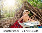 Small photo of Little blond scout boy sit with treasury map playing treasure hunt game over hut of branches