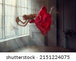 Fantasy redhead woman soars floats flies in air. Art photo levitation. Girl fairy princess reads magic book, divine light from window. Red midi dress, lon hair flutters in wind. Room classic interior