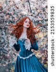 Small photo of Fantasy portrait red-haired girl romantic princess stands in spring flowering garden. Blooming green tree flowers. Long hair red lips pale skin face woman queen medieval vintage creative design dress