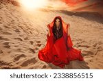 Mystery arabic woman sits in desert, gold mask veil accessories niqab burqa hide face. Fantasy girl art photo fashion model black hair head cover scarf, red long dress. Sand dunes sun light